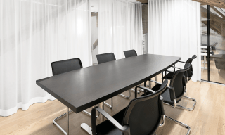 Ein kleineres Sitzungszimmer mit Parkettboden, einem langen schwarzeb Tisch, 6 Stühlen und weissen Vorhängen über verglasten Wänden.