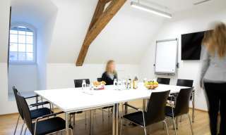 Ein helles Sitzungszimmer im Dachgeschoss mit Tageslicht, hölzernen Dachbalken, Parkett und verstellbaren Tischen und Stühlen. An der Wand sind Flipchart und Wandbildschirm angefestigt. Im Raum befinden sich zwei Personen.