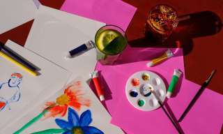 Verschiedene Malsachen, Papier und zwei Getränke sind auf einem Tisch verteilt.